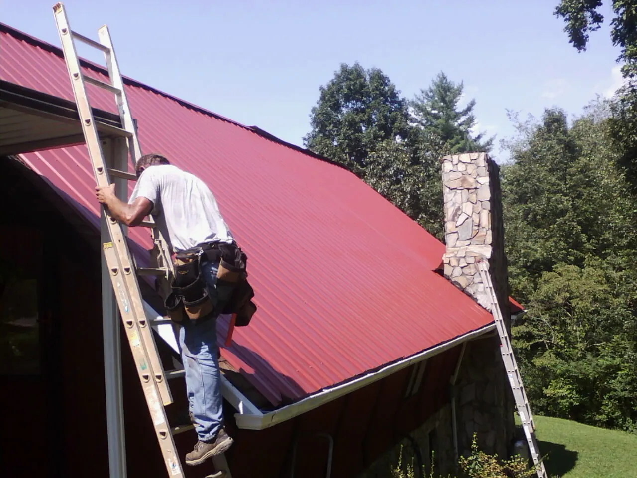 Roofer on a ladder beside a red metal roof and stone chimney — Soffit & Fascia Repair by Roofing Brothers in Apalachin
